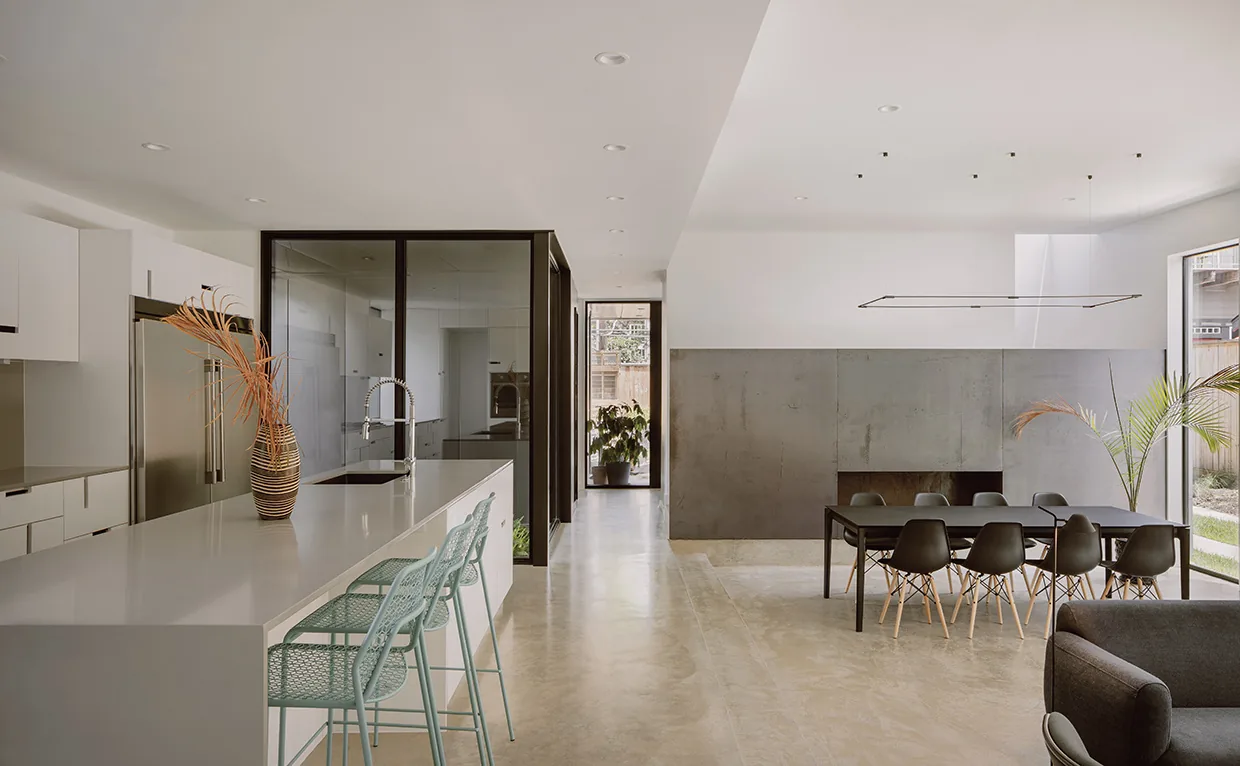 Open plan kitchen and dining area featuring concrete flooring, a white island with bar stools, and a black dining table with chairs next to floor-to-ceiling windows.