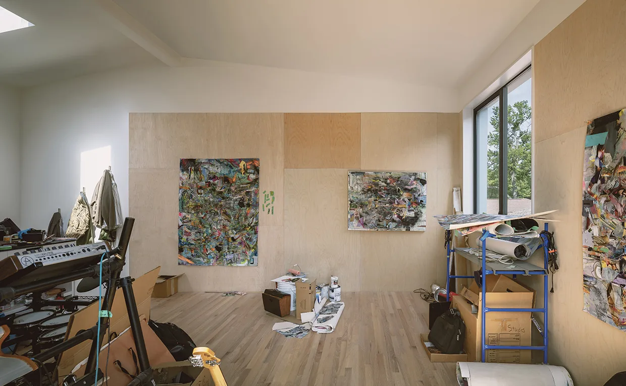 Studio space interior with wood panel walls and flooring, two large rectangular windows on the right side, and abstract paintings displayed on the walls.