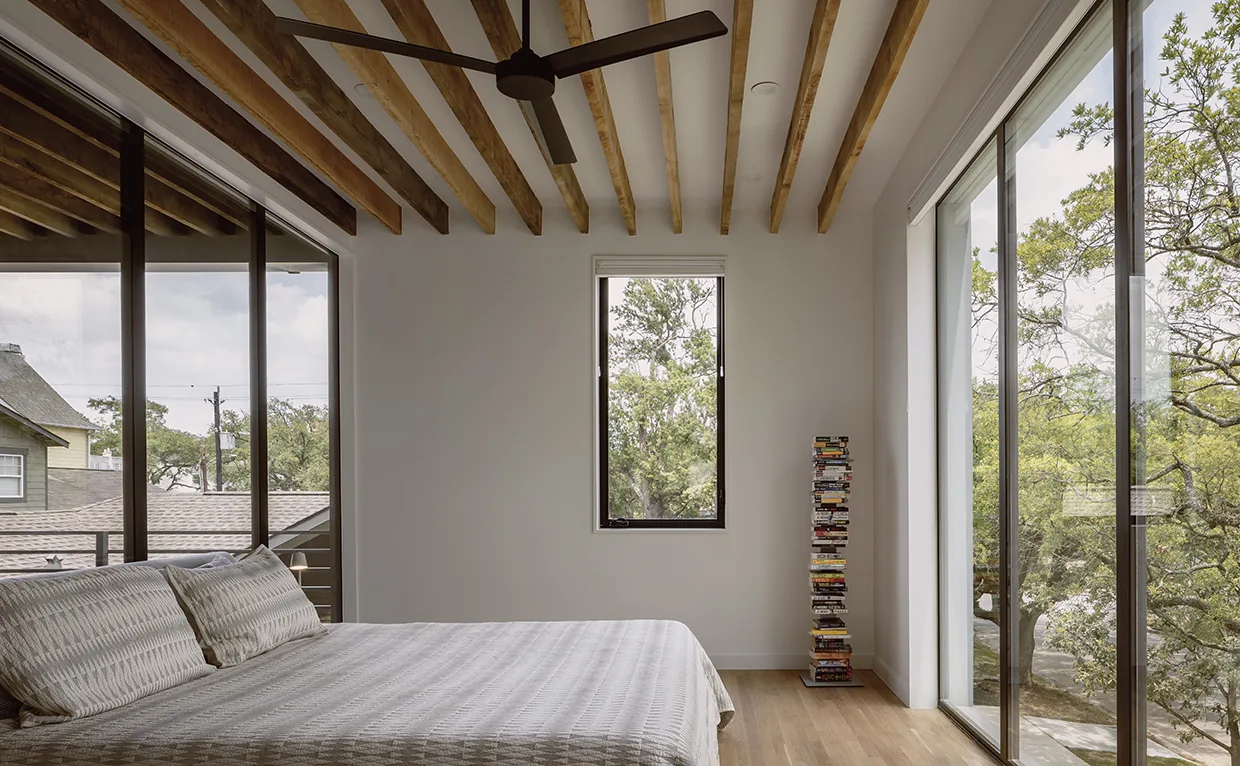 Bedroom featuring exposed wood beams, a ceiling fan, and large glass windows overlooking trees.
