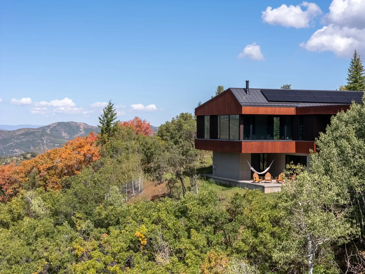 Two-story modern house with concrete base and wood-paneled upper level overlooking a mountain landscape with deciduous trees in fall colors.