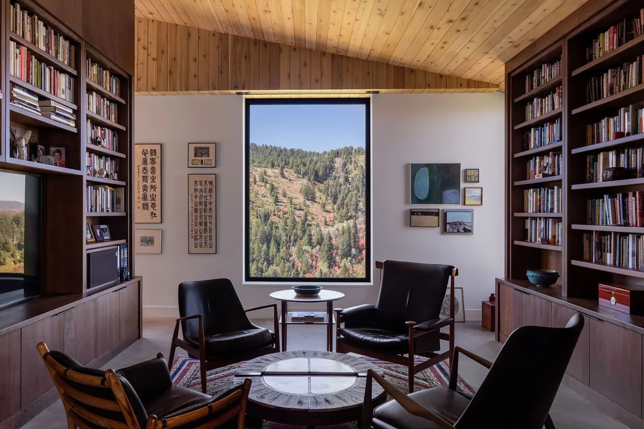 Reading room with large framed window showcasing forested hillside view, wood-paneled ceiling, and built-in bookshelves with books and decor.