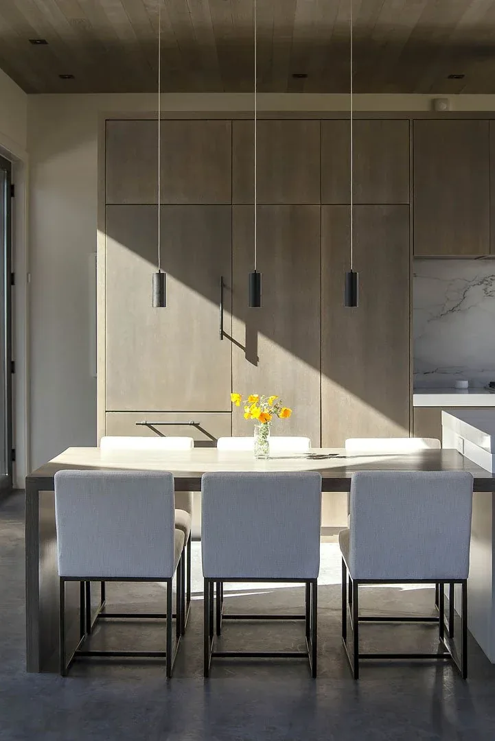 Dining area with six white upholstered chairs and modern cabinetry, featuring three black pendant lights above the table.