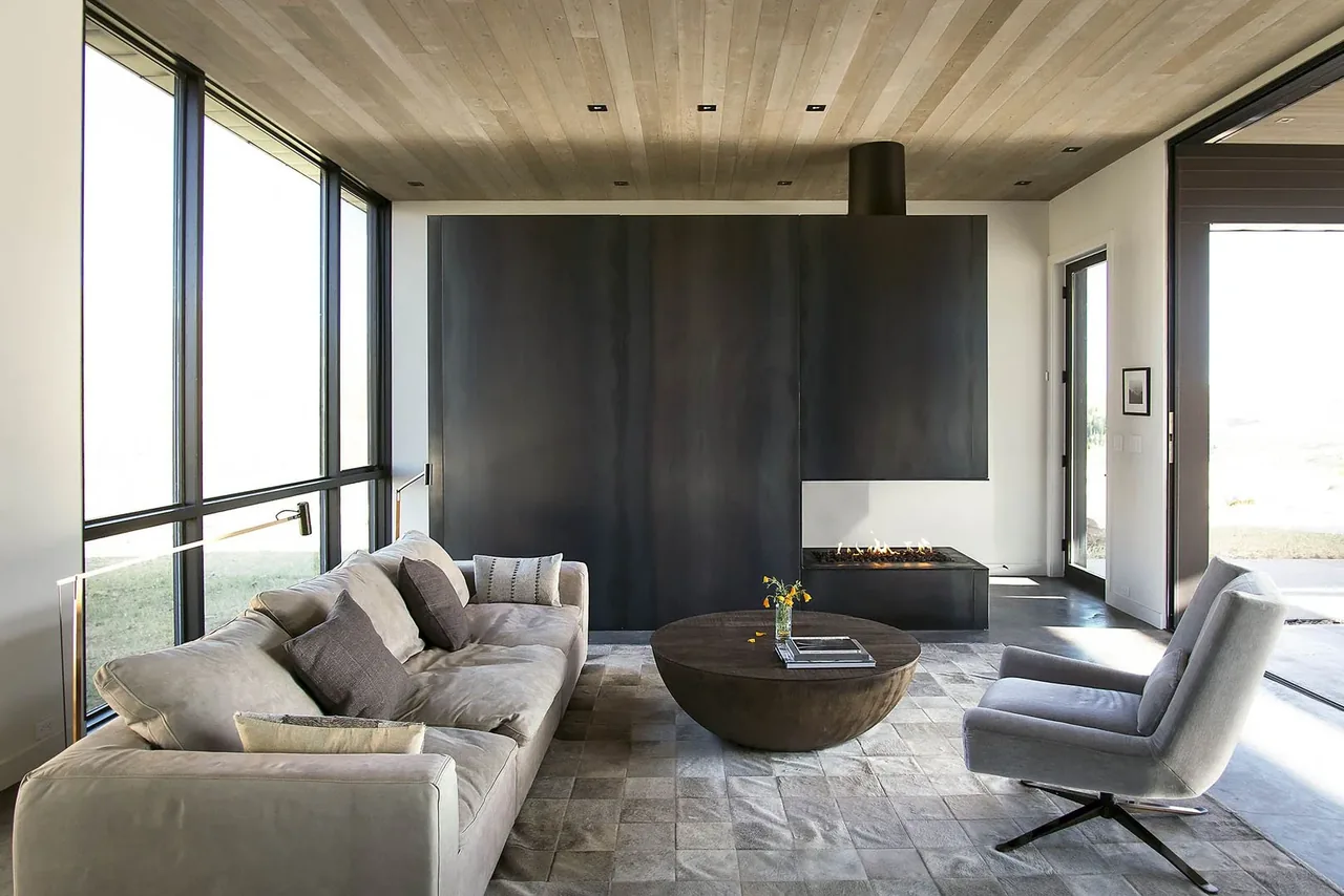 Living room with a large black fireplace wall, floor-to-ceiling black window frames, and modern furniture.