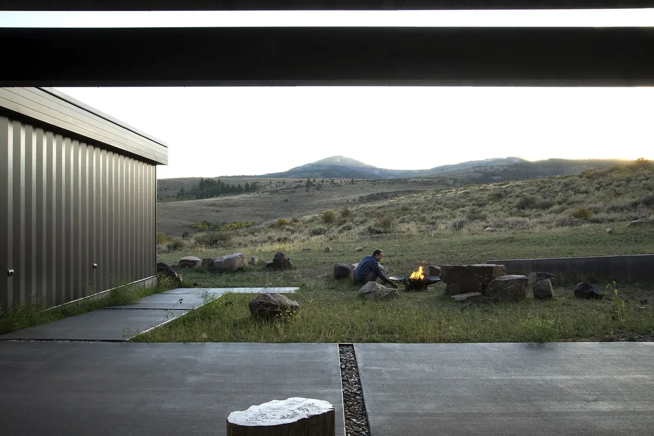 Covered patio adjacent to modern metal building with stone fire pit and rural hillside landscape.