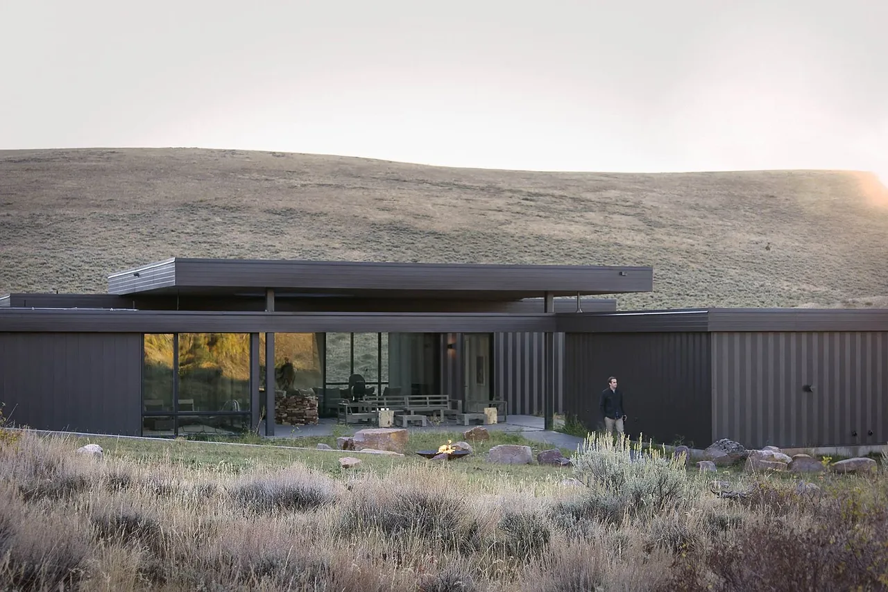 Modern single-story house with floor-to-ceiling glazing in a rural hillside setting with dry grass and stone fire pit.