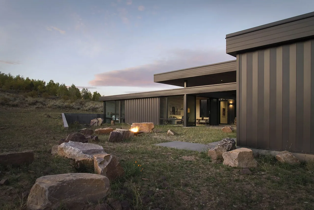 A modern house with metal siding features a stone fire pit and large glass walls overlooking a rural hillside.