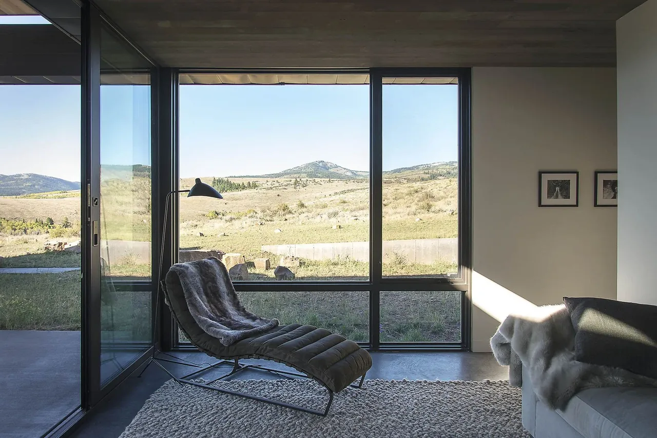 Living room with floor-to-ceiling black window frames overlooking open hillside landscape.