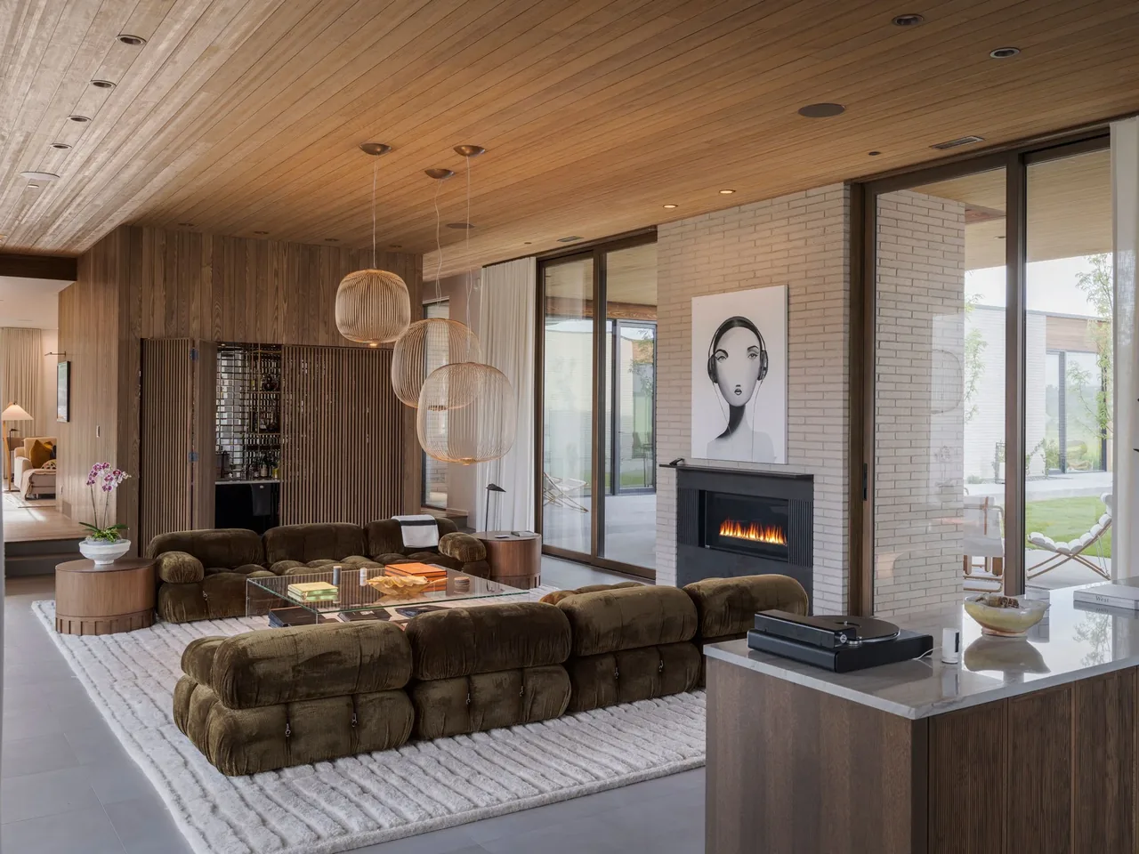 Interior view of a modern living room featuring a brown sectional sofa, brick fireplace, and floor-to-ceiling glass doors.