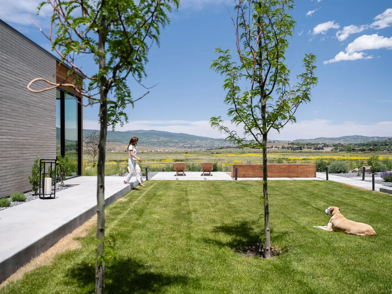 Exterior view of a modern house showing brick wall, concrete walkway, grassy lawn, and outdoor seating area in the distance.