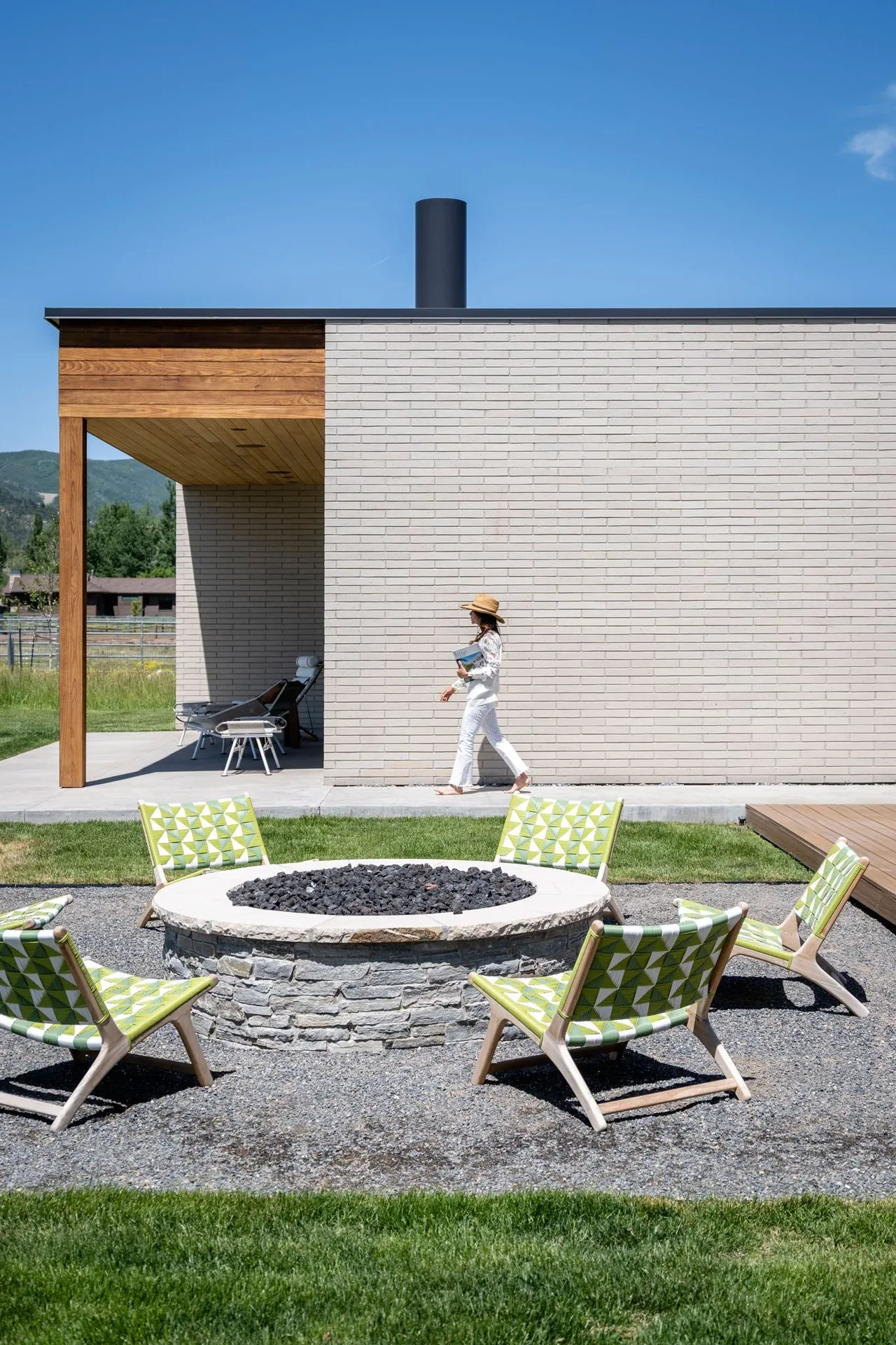 Outdoor area with a stone fire pit, surrounded by low chairs with green geometric patterned fabric, adjacent to a modern brick and wood house exterior.