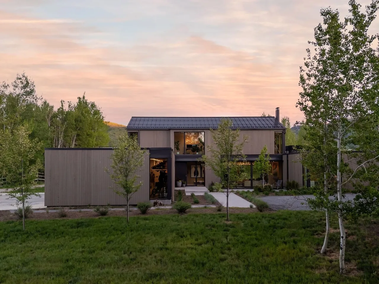 Modern two-story house with wood siding and large windows set against a natural backdrop at sunset.