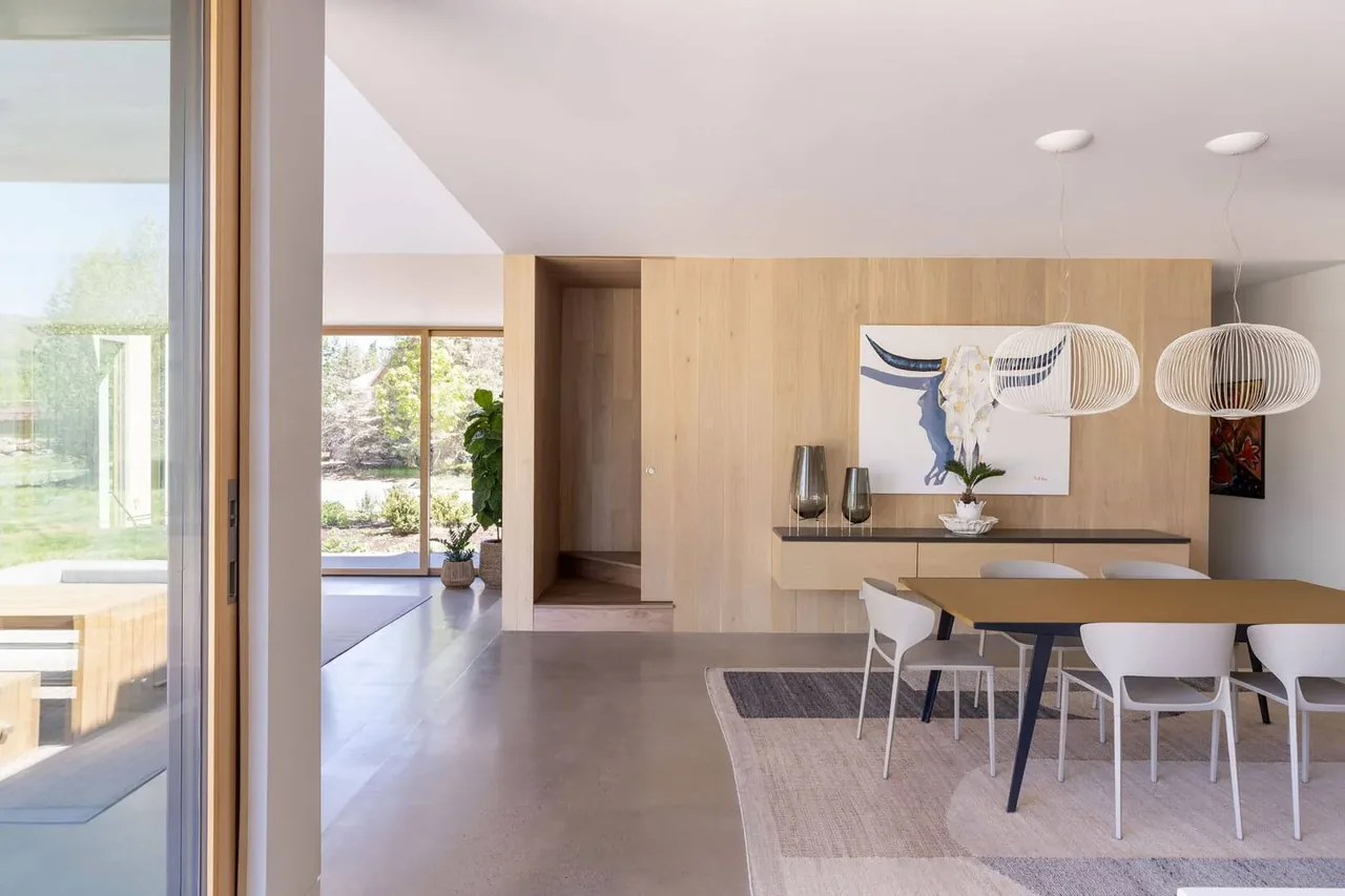 Dining space features vertical light wood paneling, modern pendant lights, and a table with white chairs, next to a large sliding glass door.