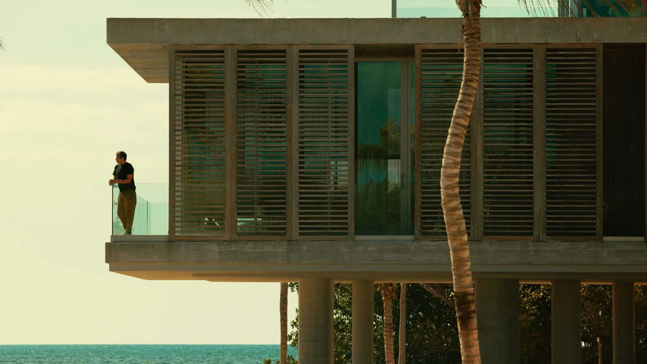 Cantilevered concrete upper floor with vertical wooden slat sunshades and glass balcony overlooking a waterfront landscape.
