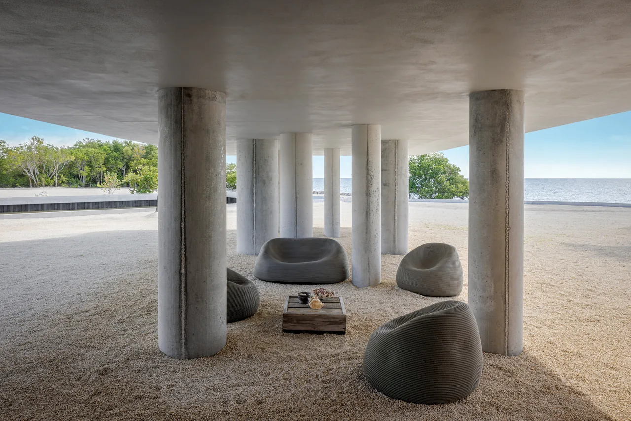 Concrete columns support a cantilevered overhang above a gravel-covered outdoor seating area by the water.