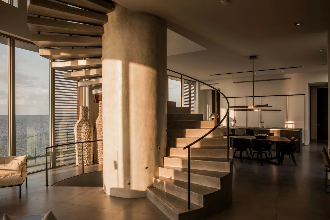 Curved concrete staircase with metal railing in a modern residence interior overlooking the ocean through floor-to-ceiling windows.
