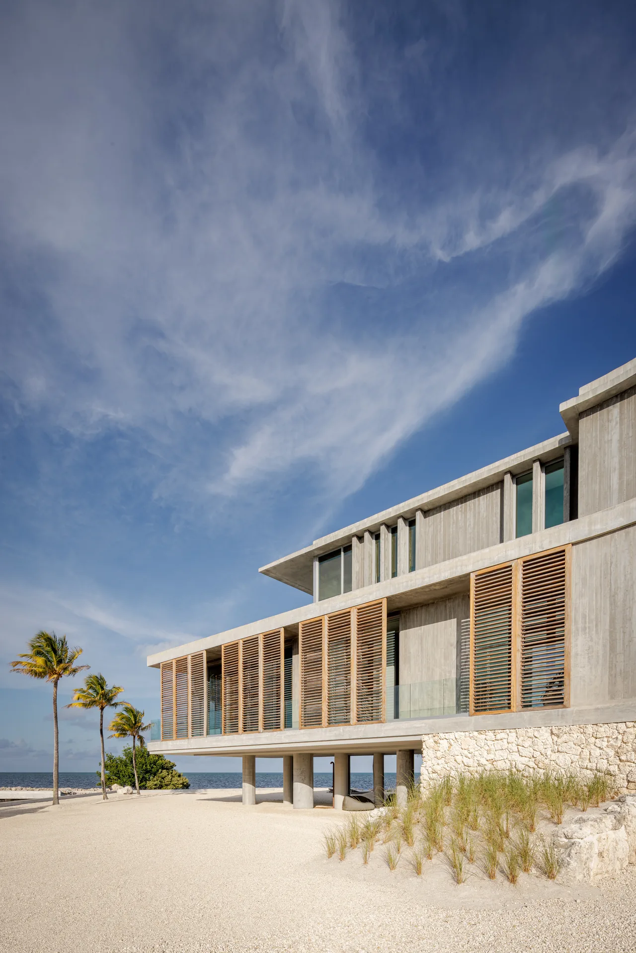 Concrete residence with cantilevered upper levels featuring vertical wood slatted screens and glass balconies facing the water