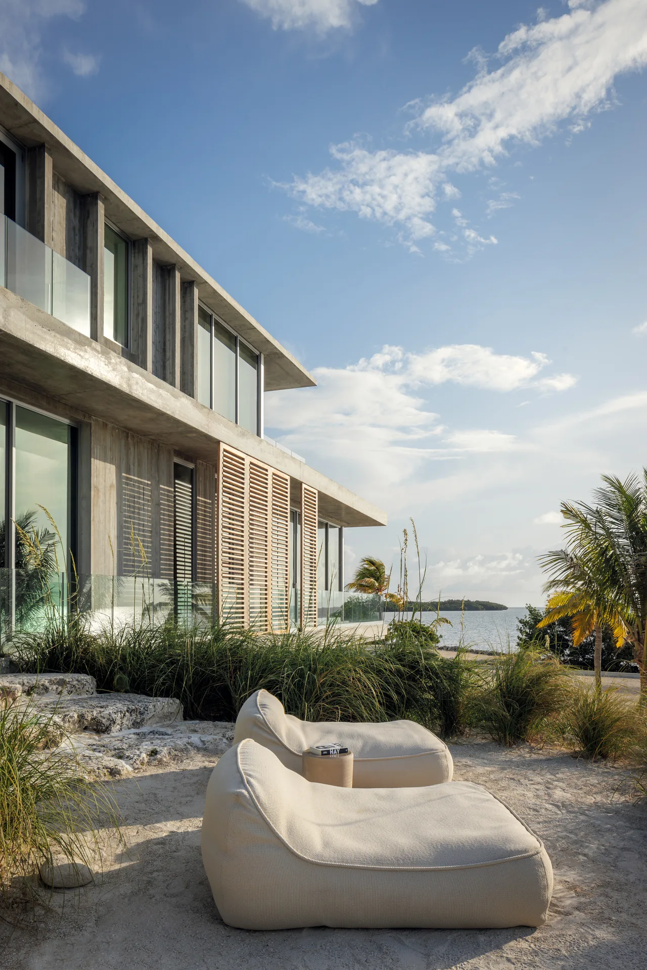 Exterior side view of a modern concrete residence featuring cantilevered design, wood slatted shades, and coastal landscaping.