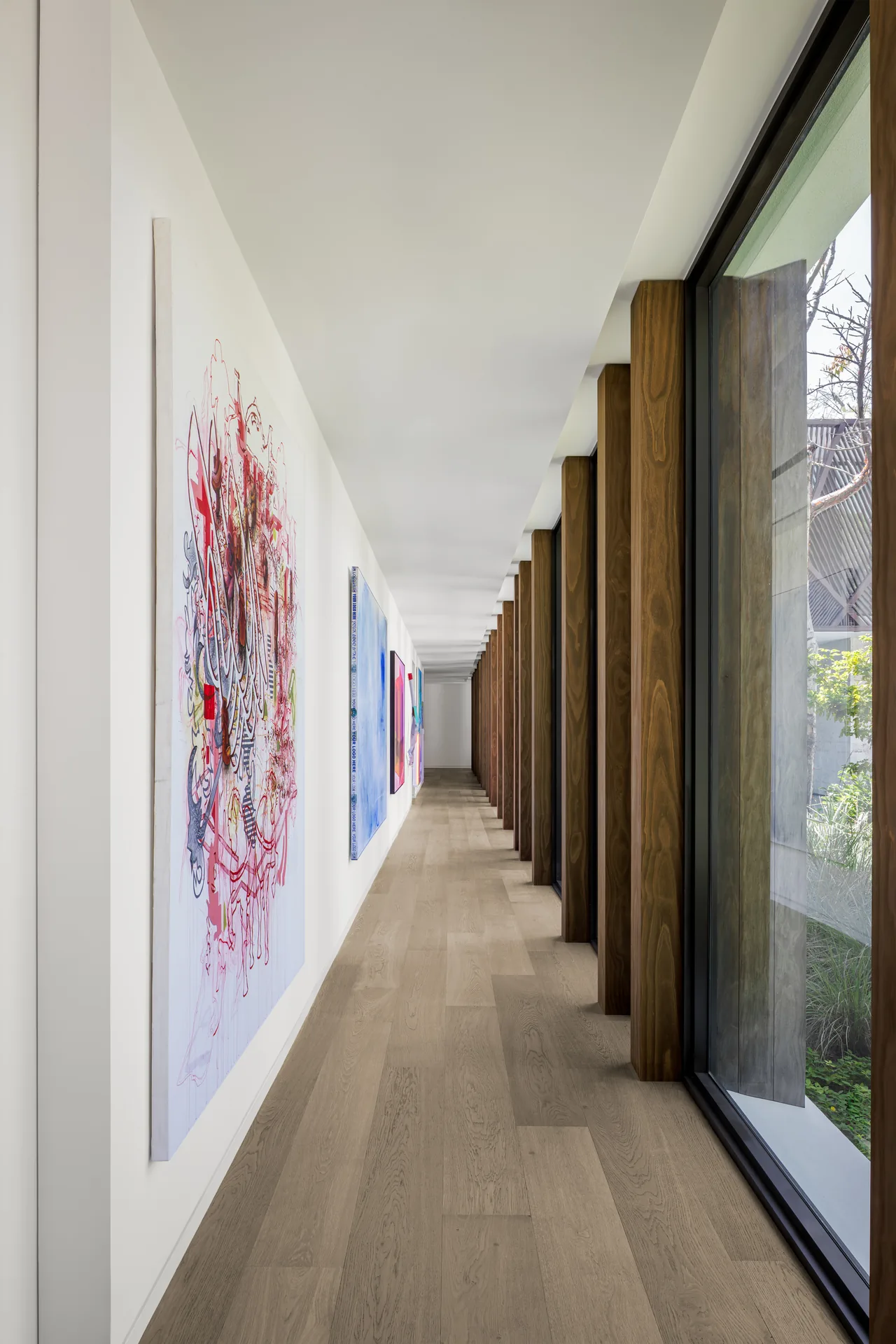 Hallway with light wood floor, white walls decorated with paintings, and floor-to-ceiling windows framed by vertical wood supports.