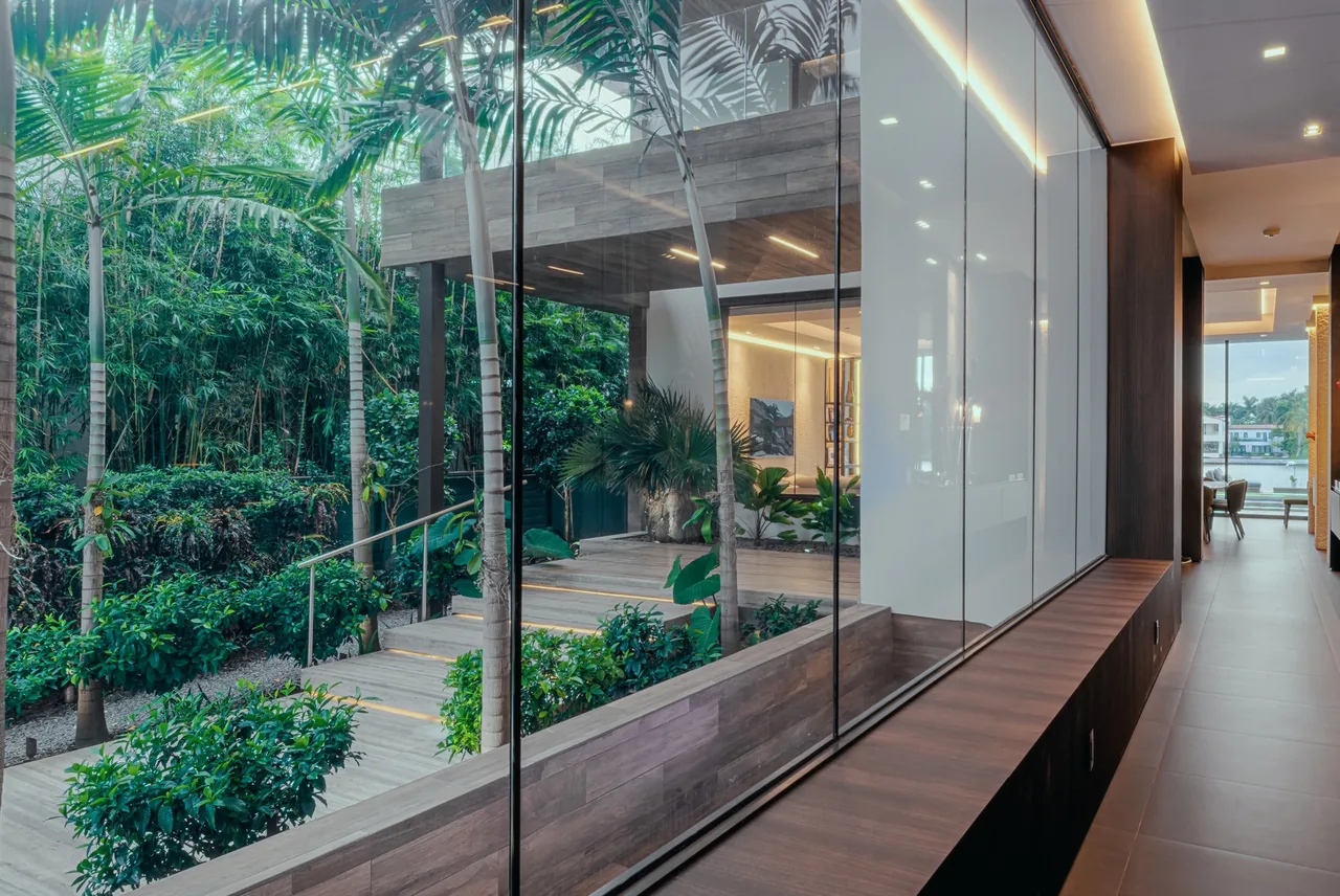 Floor-to-ceiling glass separates the interior hallway from a lush outdoor garden with tropical landscaping and wooden steps.
