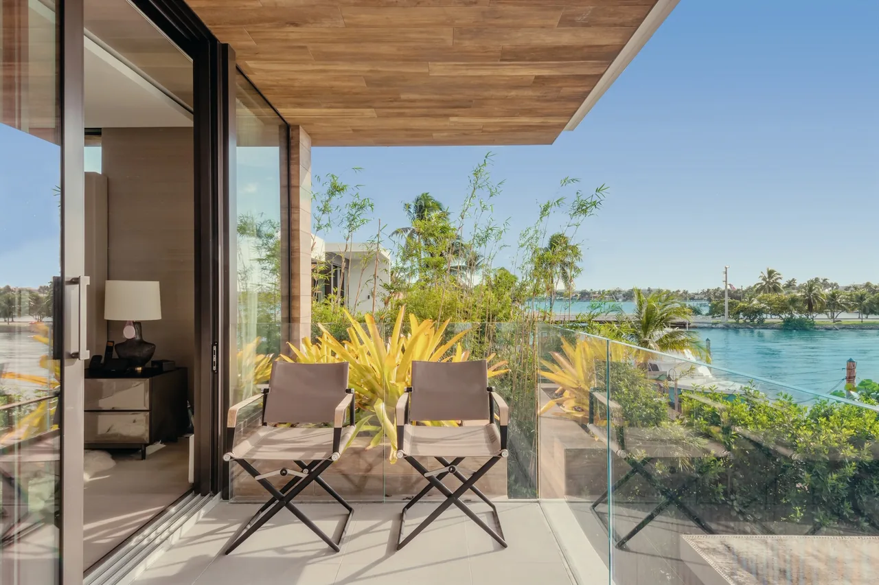 Balcony featuring folding chairs, glass balustrade, and tropical landscaping with water views in the Net Zero House.