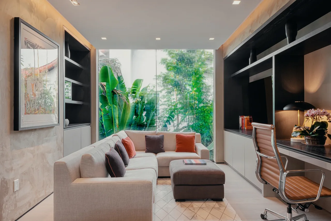 Living area features a beige sectional sofa, built-in black shelving, and floor-to-ceiling glazing with tropical landscaping views.