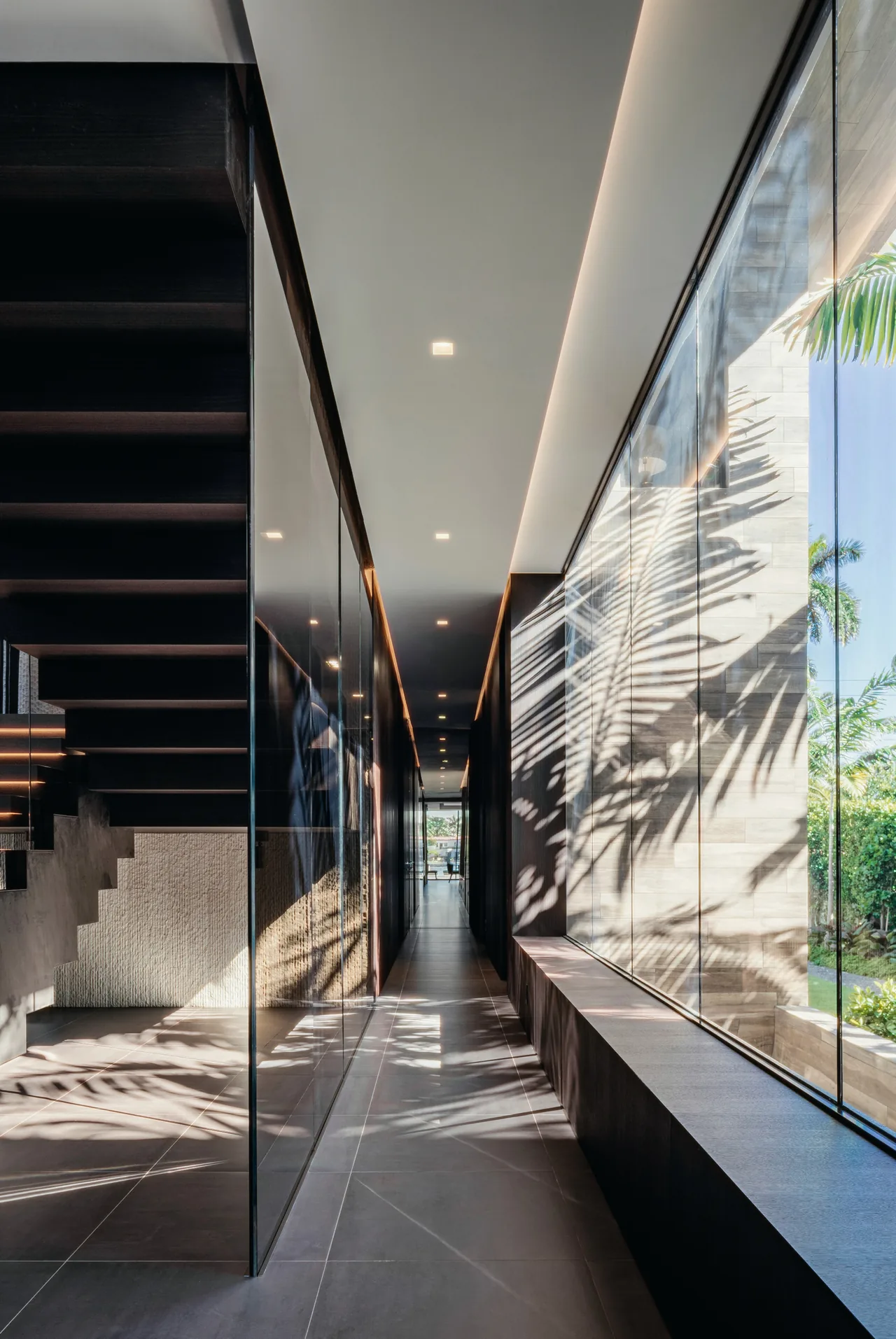 A hallway with a glass wall casting shadows of palm leaves, featuring a floating dark wood staircase and ambient ceiling lighting.