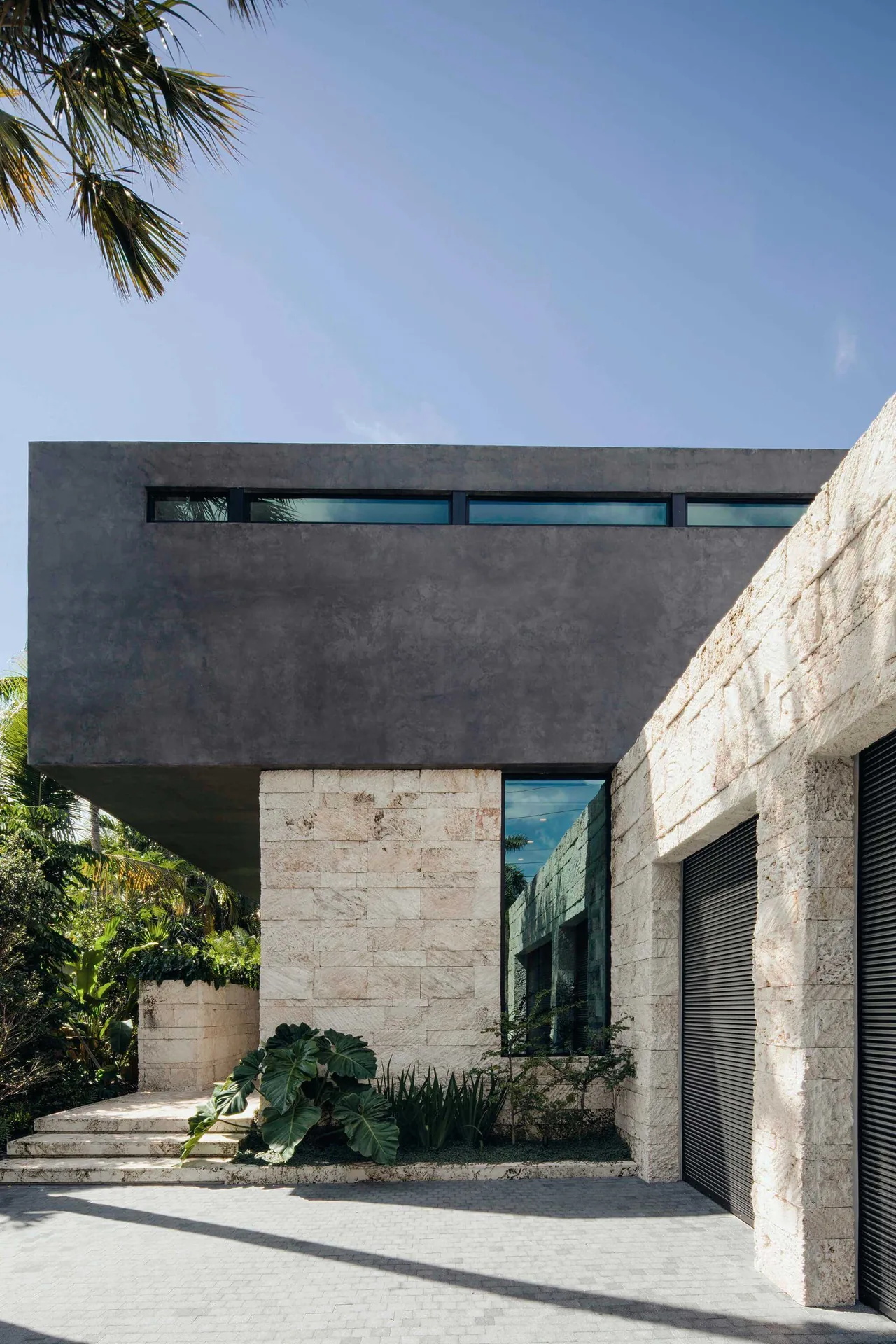 Janoura Residence exterior features a cantilevered concrete volume above natural stone walls with reflective strip windows.