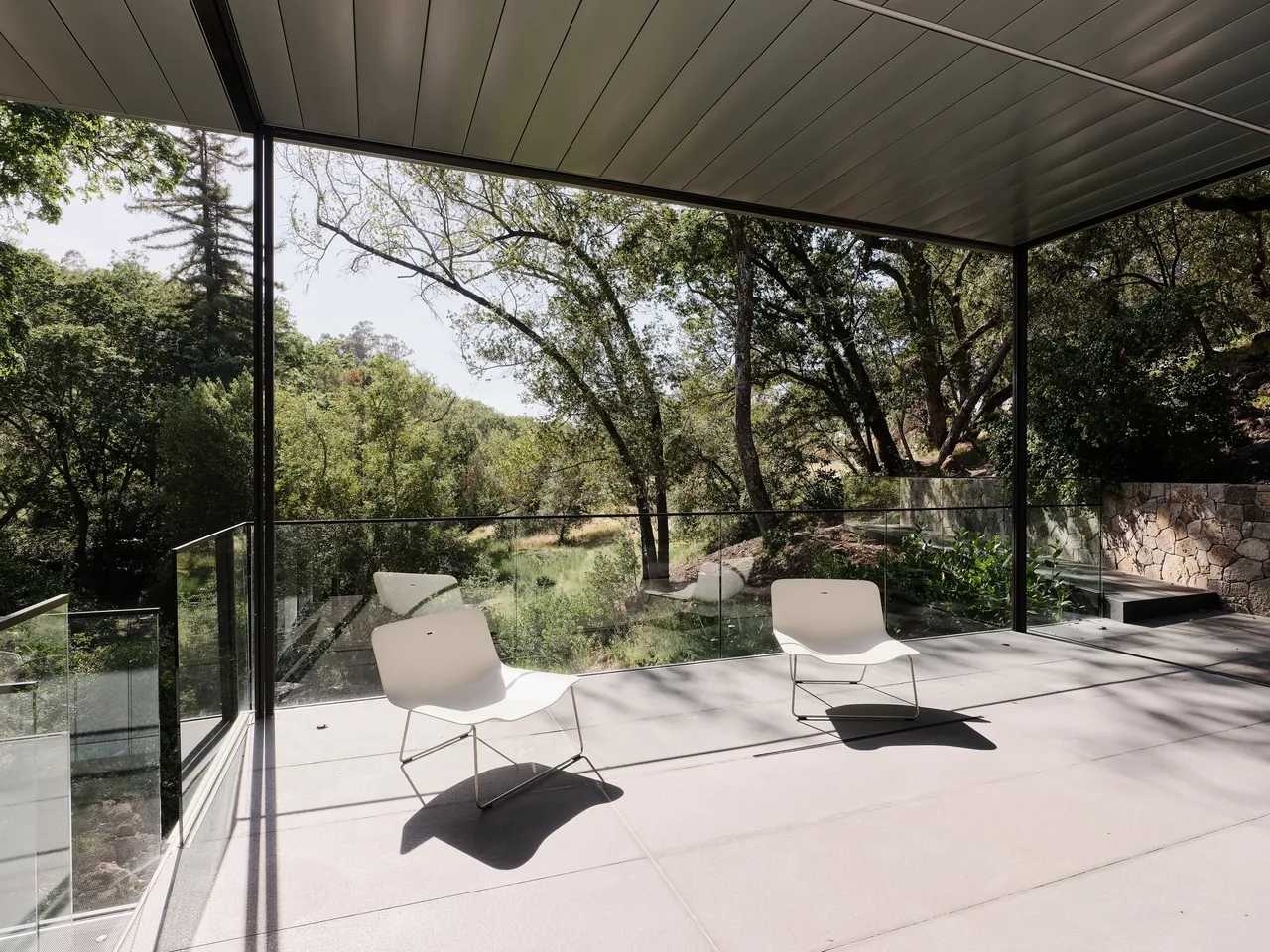 Patio area of the Suspension House with glass railing and minimalist white chairs under a metal roof.