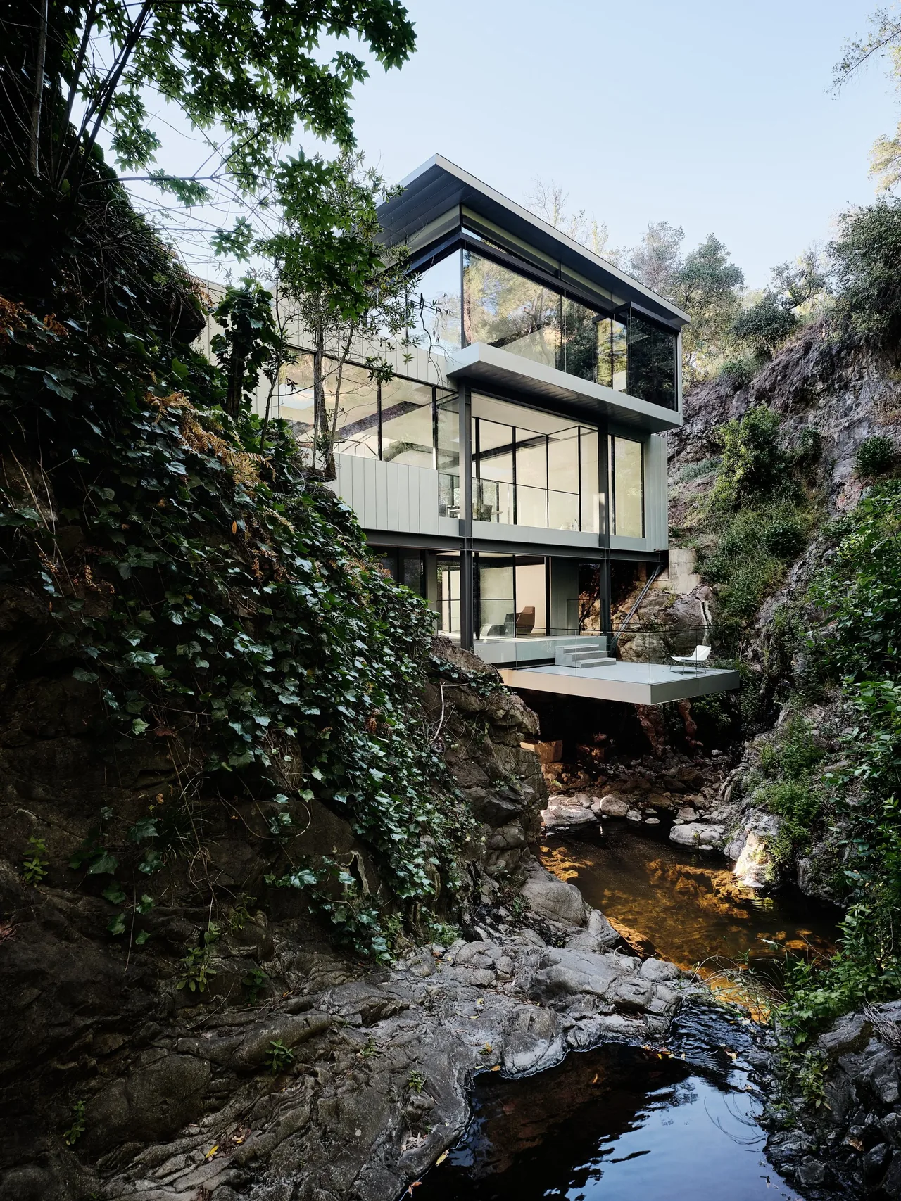 The Suspension House features extensive glass walls and a cantilevered deck extending over a rocky stream.