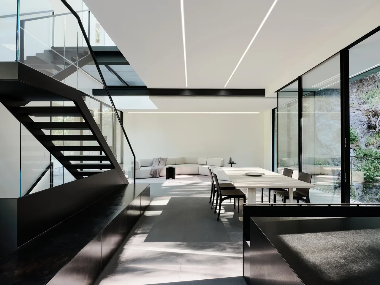 Open-plan living and dining room in the Suspension House by Fougeron Architecture features a black steel floating staircase and expansive glass walls.