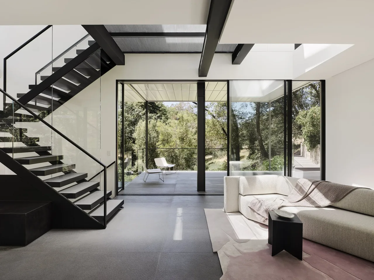 Interior view of the living room featuring a black steel staircase, floor-to-ceiling glass, and a balcony overlooking wooded hills.