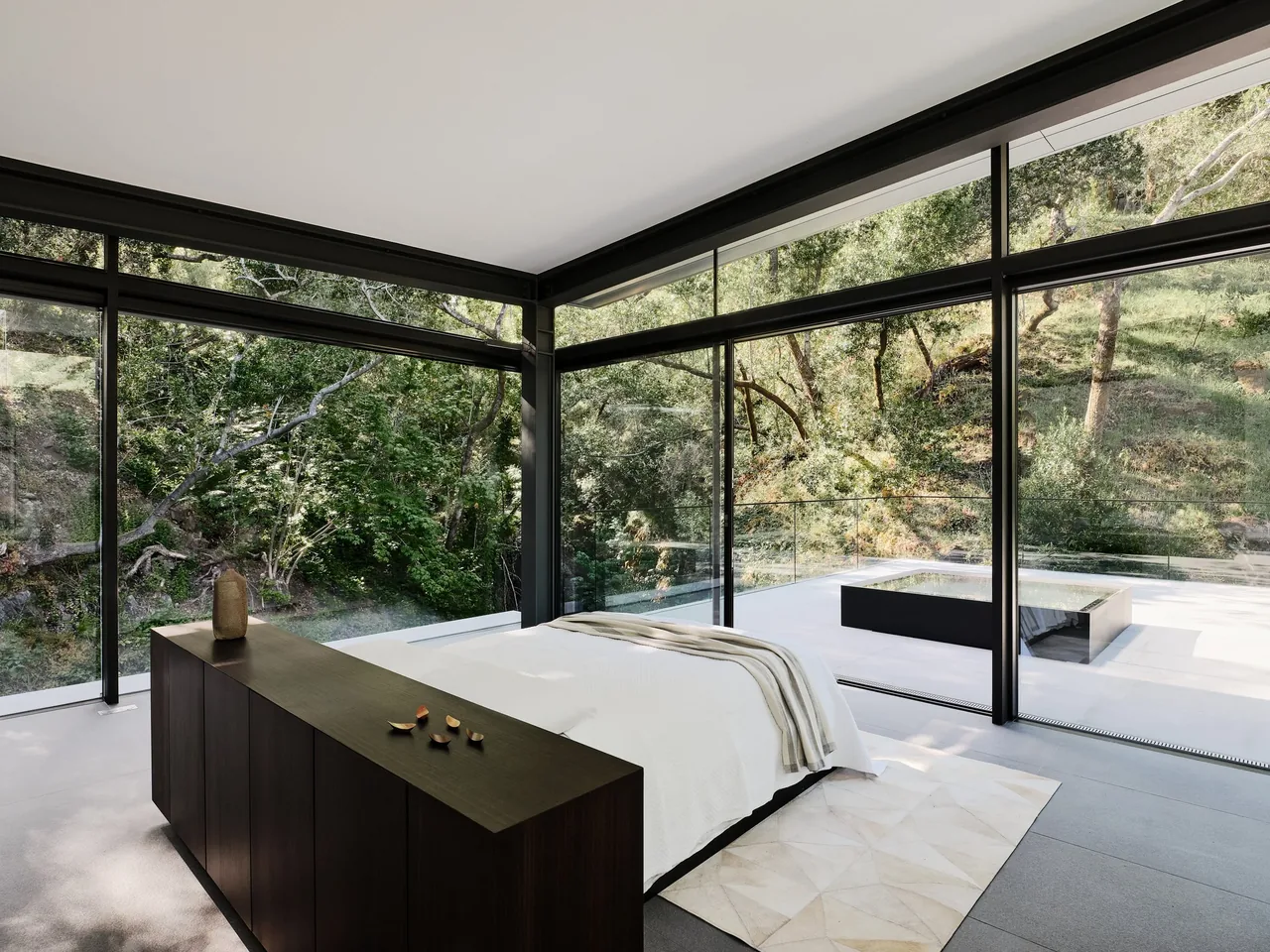 The bedroom features floor-to-ceiling glazing with black window frames and a view of the wooded hillside.
