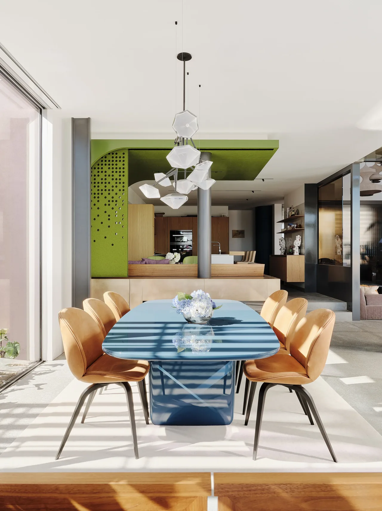 Dining room features a blue modern table with six tan leather chairs and a geometric pendant light, adjacent to large windows causing shadow patterns.