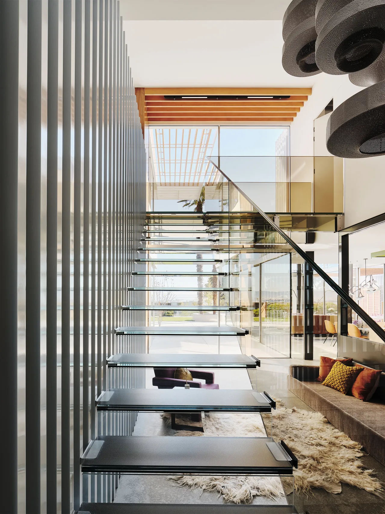 This modern interior features a floating glass staircase next to a cozy living area with a sheepskin rug and sofa with orange pillows.