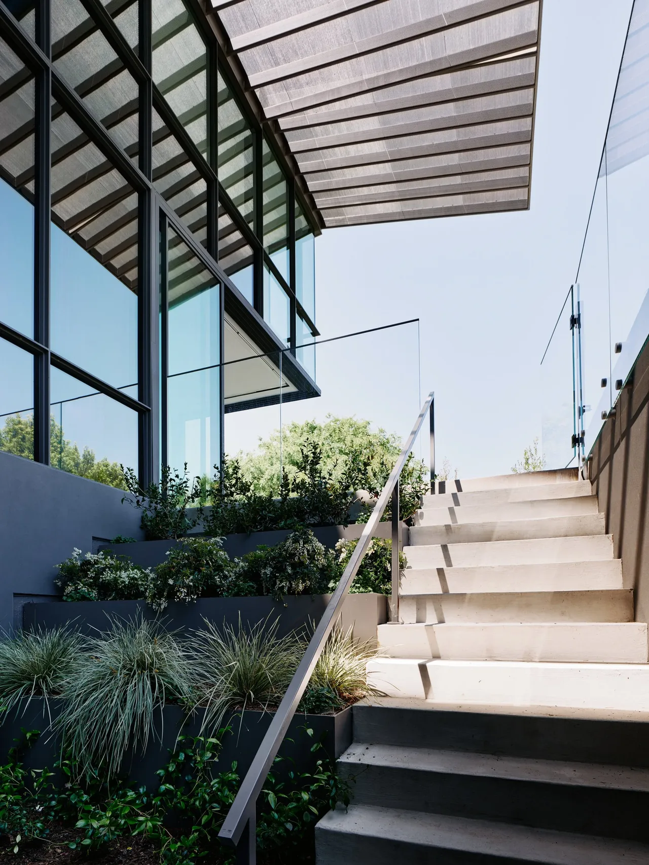Concrete exterior stairs next to large corner windows and a metal mesh pergola casting shadows.