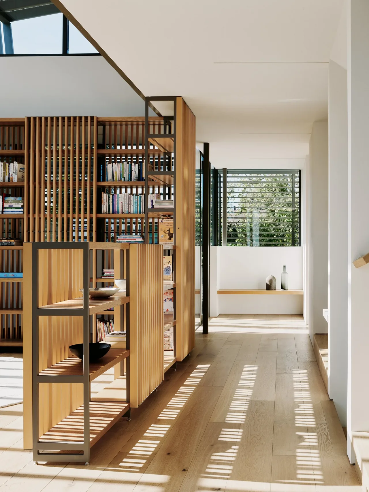 Sunlight casts striped shadows from wooden slatted shelves and window horizontal slats on the light wood floor in a modern hallway.