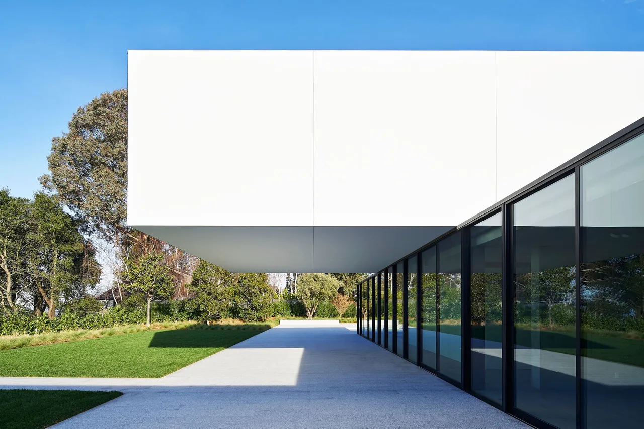 Cantilevered white upper volume over a concrete pathway with black window frames and landscaped green lawn in the Oz Residence.