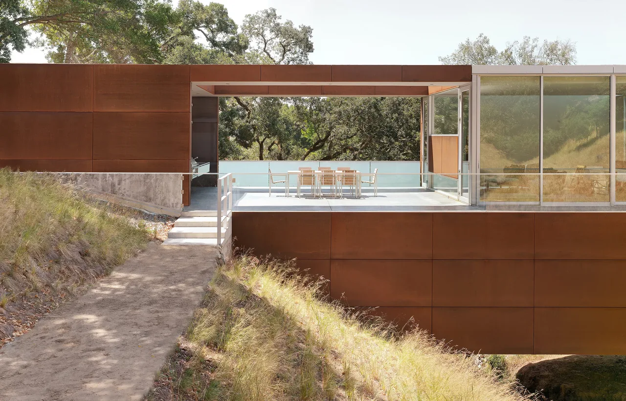 Exterior front view of Bridge House showing the steel-clad elevated structure with floor-to-ceiling glazing and a terrace dining area.