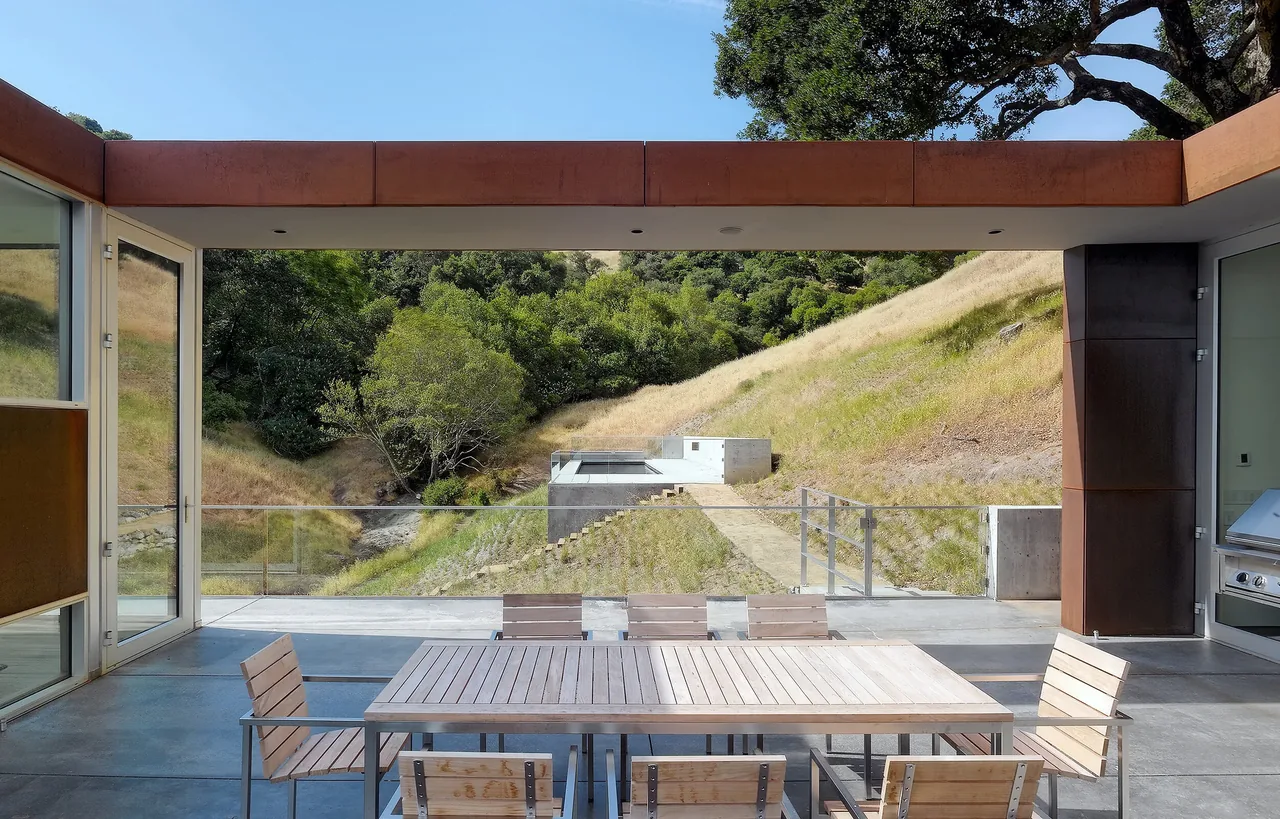 View through an open exterior dining space featuring steel framing and rust-colored flat roof, overlooking a natural hillside and concrete landscape elements.