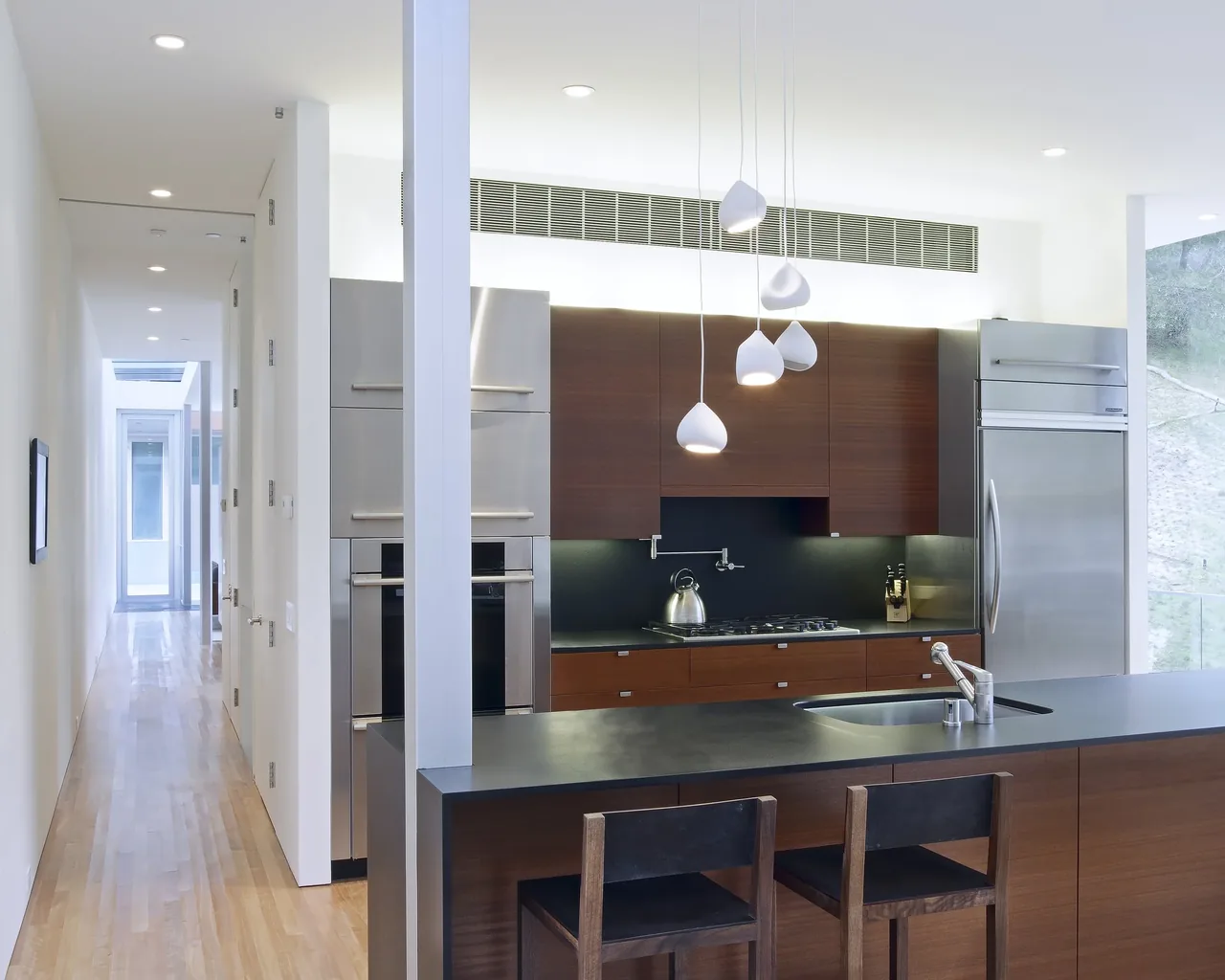 Kitchen space featuring dark wood cabinetry, a black countertop island with built-in sink, stainless steel refrigerator and ovens, and pendant lighting.