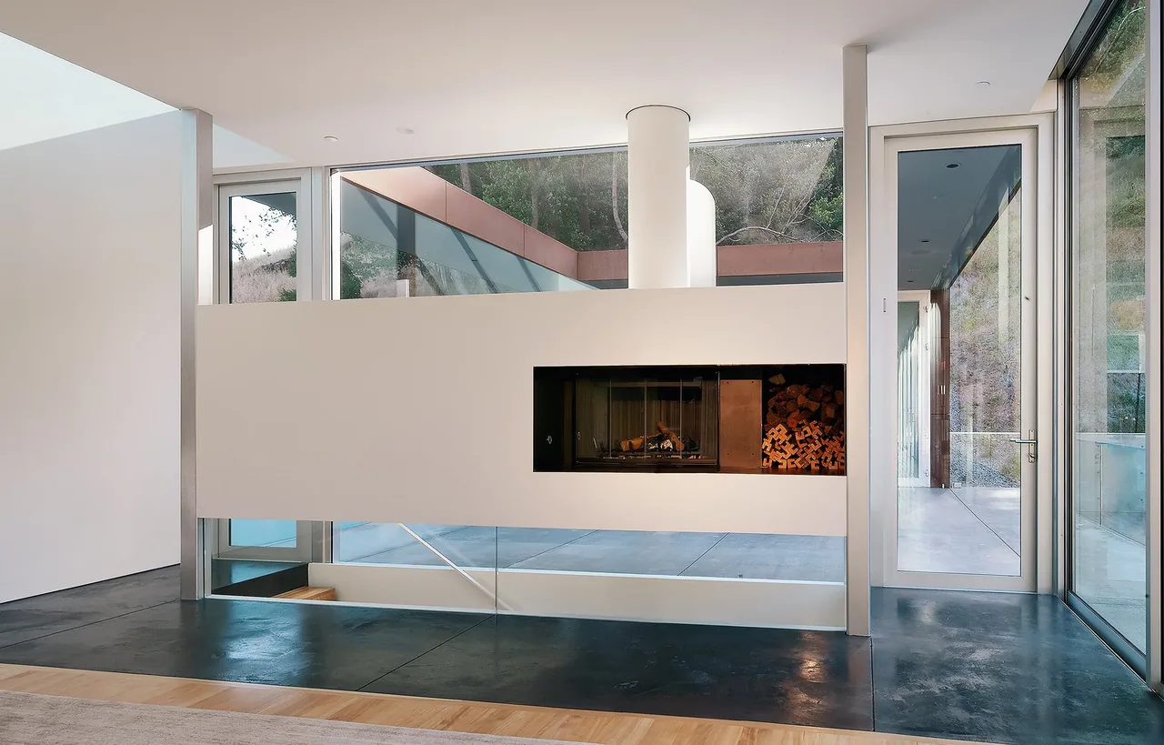 Interior view of a modern room with a built-in gas fireplace and wood storage integrated into a white partition wall, surrounded by floor-to-ceiling glass walls and doors.