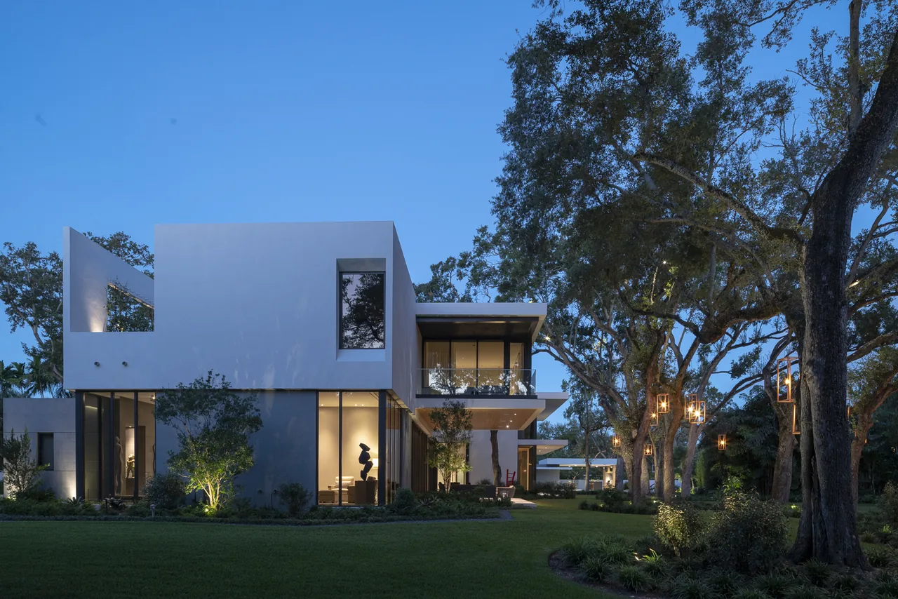 Exterior rear view of a modern two-story residence with large windows and integrated landscape lighting around trees and lawn at dusk.