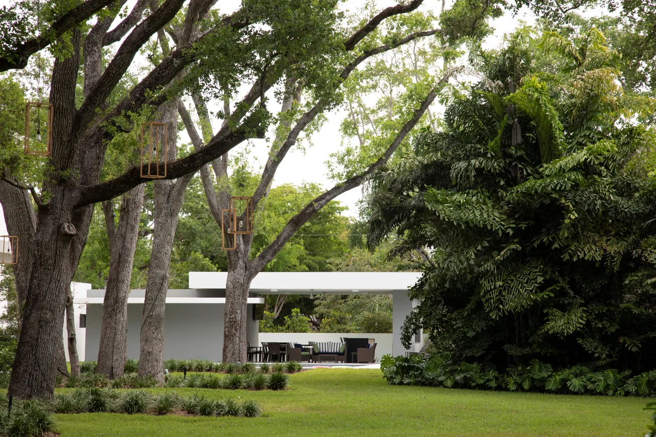 A minimalist white pavilion in a landscaped yard with mature trees and planted garden beds.