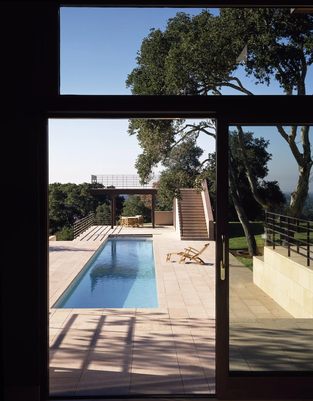 Outdoor view from inside shows rectangular pool, stone terrace, and metal railing balcony with stairs leading up.