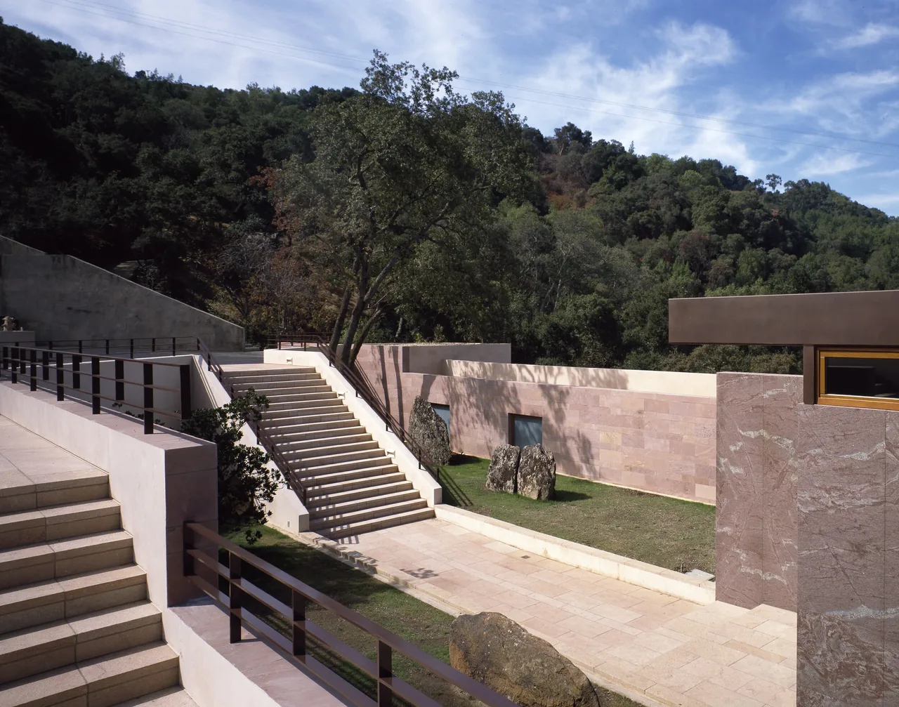 Exterior stairs and ramp with metal railings next to stone walls and a landscaped area with rocks and trees.