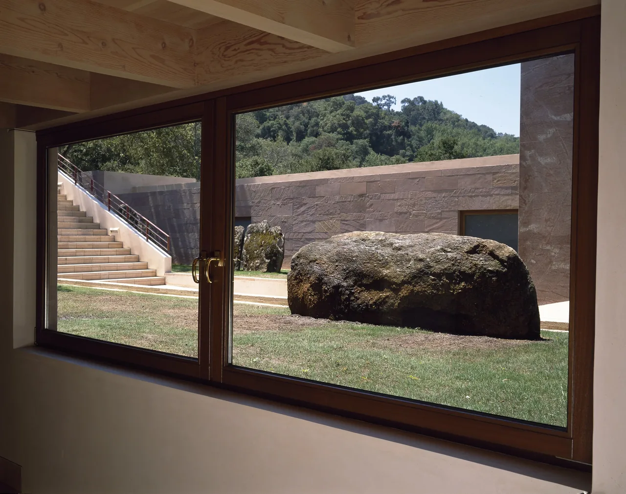 Courtyard view through large wood window features stone wall, stone steps, and a boulder with forested hills beyond.