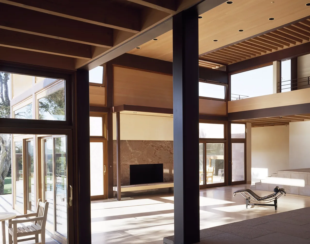 Di Napoli Residence living room with exposed beams, large windows, and a stone fireplace by Stanly Saitowitz | Natoma Architects.