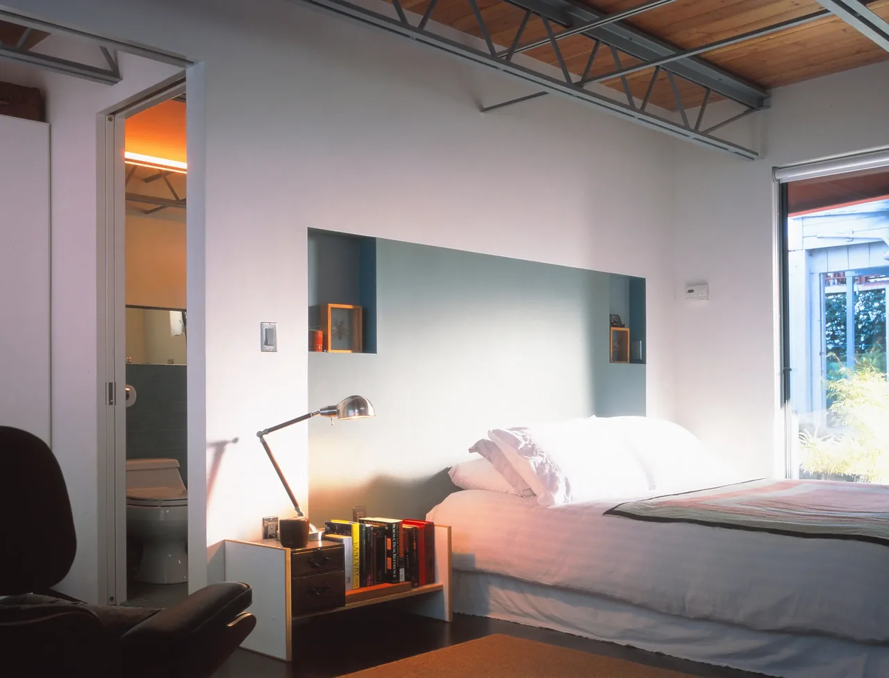 Bedroom features a white bed against a blue recessed headboard wall, an industrial ceiling with exposed steel trusses, and a sliding door to the bathroom.