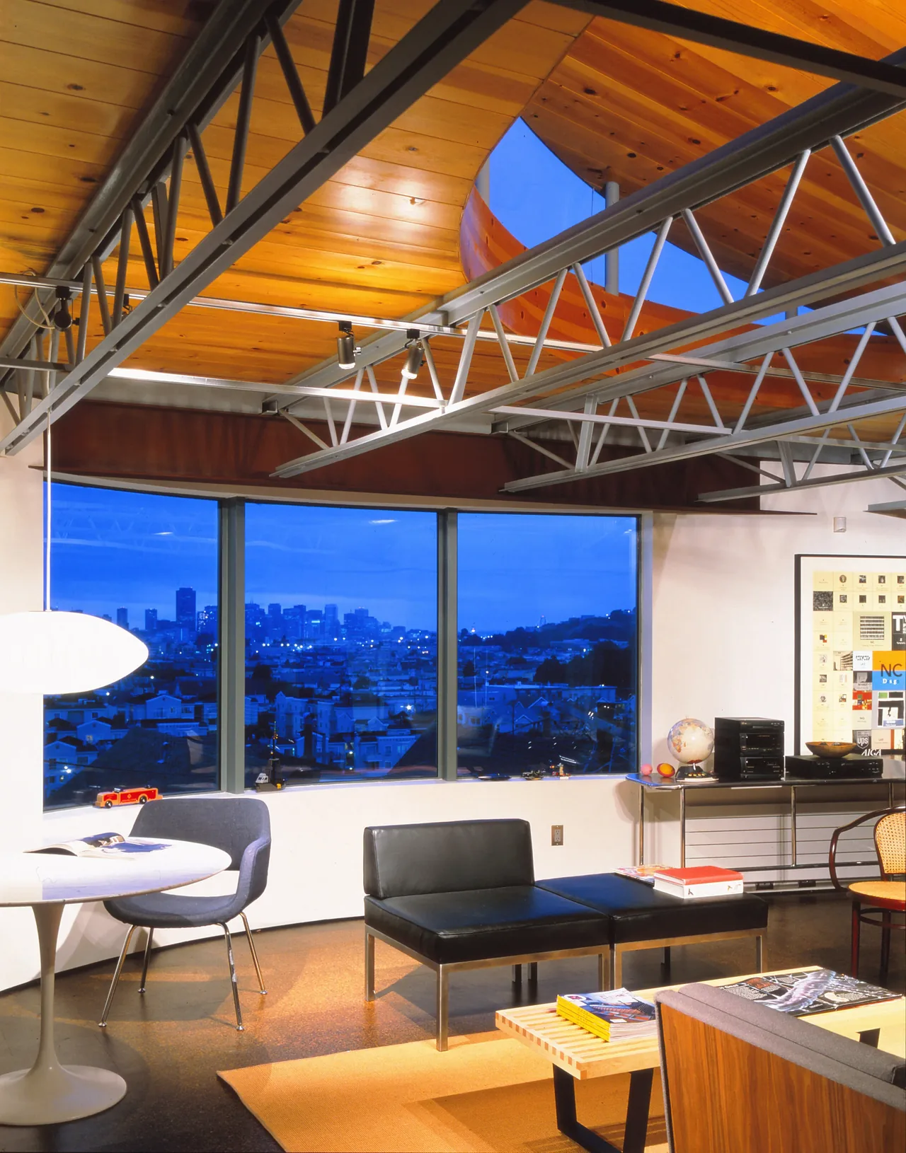 Barnes Residence living area features a curved wood ceiling with exposed metal trusses and large picture windows framing a city view at dusk.