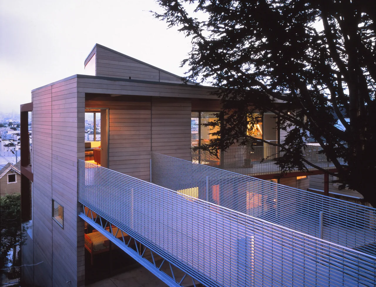 The Barnes Residence features wood siding and glowing windows with a metal balcony outside rooms on a sloped site.